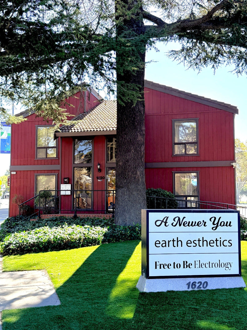 A red two-story building with a large tree in front, alongside a white sign that reads 'A Newer You earth esthetics Free to Be Electrology 1620' on a green lawn.