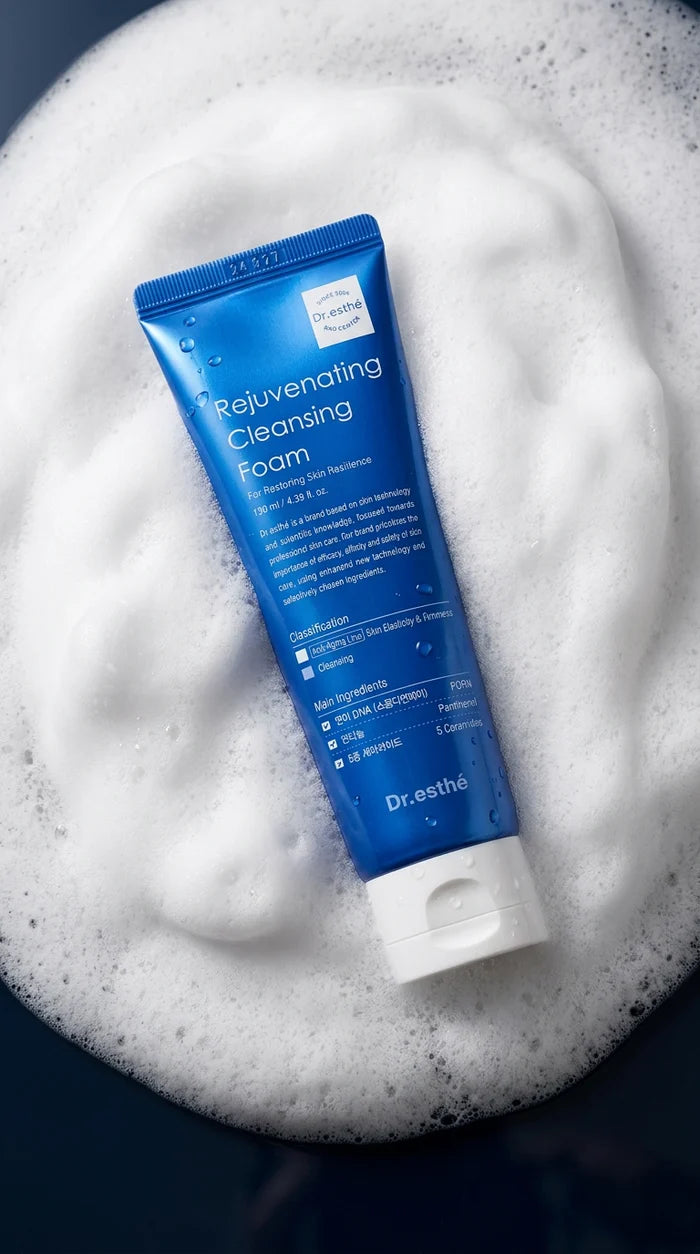 A blue tube of Dr. Estha Rejuvenating Cleansing Foam resting on a bed of white foam against a dark background.